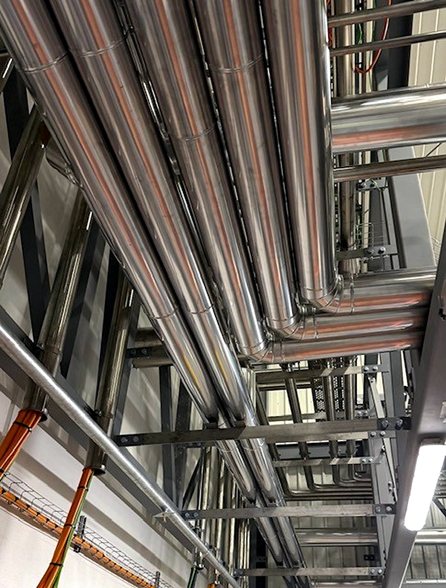 Metal cladding on overhead insulated pipe bends above industrial machinery in a factory
